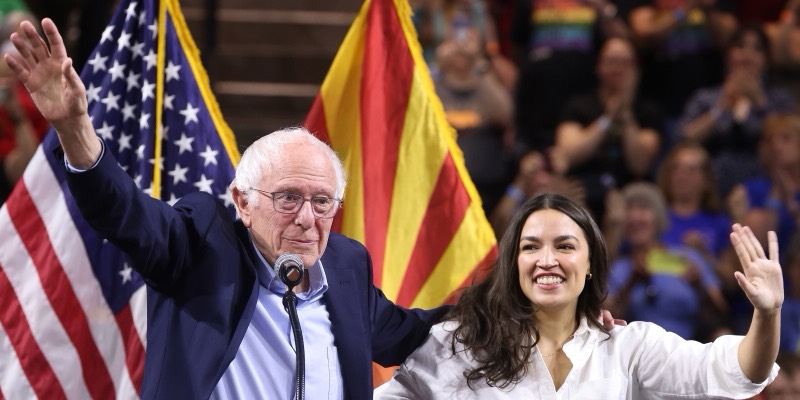 Bernie Sanders and Alexandria Ocasio-Cortez waving on stage in front of US and Arizona flags, with a crowd behind them