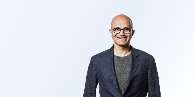 Satya Nadella, smiling, in glasses, a navy jacket and grey T-shirt, against a light blue-white background