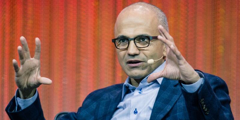 Satya Nadella, in dark-rimmed glasses, a navy checked jacket and light blue shirt, gesturing with both hands as he speaks on stage in front of a red curtain