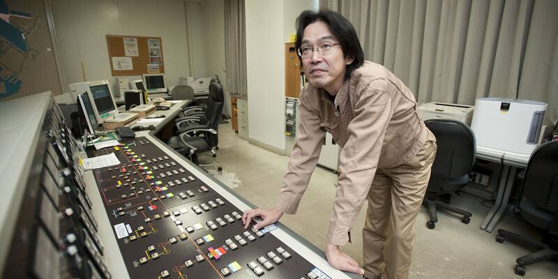 A man in a tan work uniform leans over a long control console covered in switches and a coloured electrical circuit diagram in a power control room