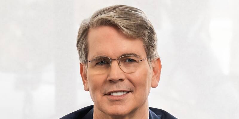 Scott Bessent, smiling, with grey swept-back hair and wire-rimmed glasses, wearing a navy suit and light blue shirt against a pale background