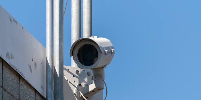 A white outdoor security camera on a metal bracket, mounted beside steel poles against a clear blue sky.