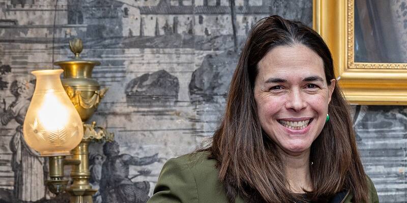 Dartmouth President Sian Beilock smiling in an olive jacket in front of a grey mural wallpaper and a brass lamp