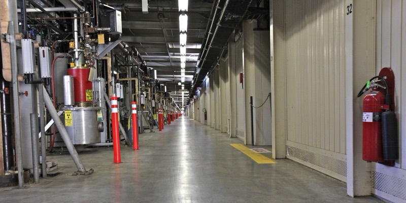 A long corridor lined with klystron tubes, pipes and red safety posts stretching to a vanishing point, with fire extinguishers on the wall