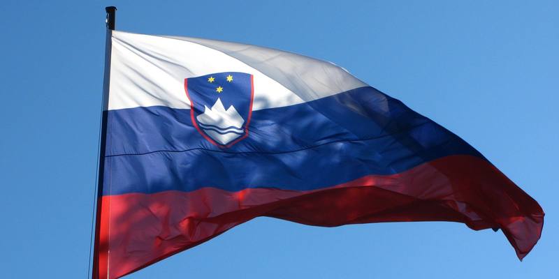 The white, blue and red flag of Slovenia, with its coat of arms, flying from a pole against a clear blue sky.