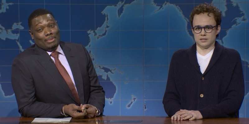 Michael Che in a suit and Jane Wickline in curly hair, round glasses and a navy cardigan sit at the Weekend Update desk in front of a blue world map