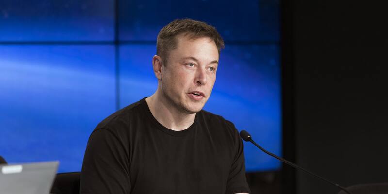 Elon Musk in a black T-shirt speaking into a microphone at a press conference table in front of a blue screen