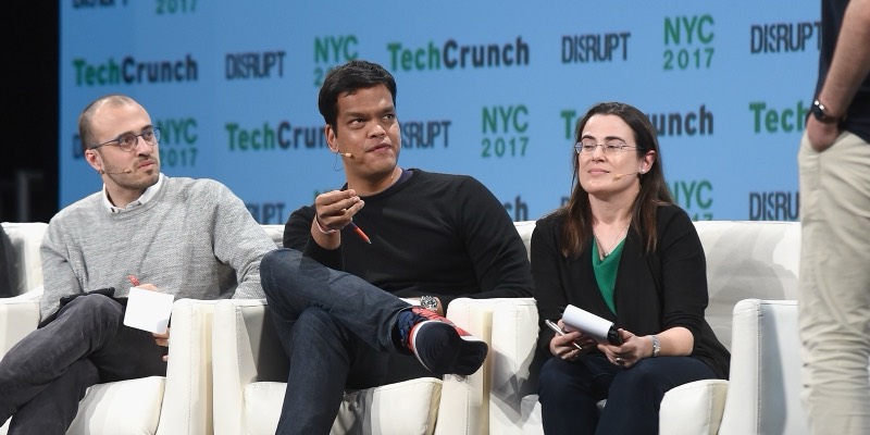 Sriram Krishnan, in a black sweater, gestures with a pen while seated between two other panellists on white chairs in front of a TechCrunch Disrupt backdrop