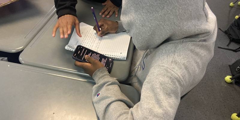 A student in a grey hoodie writes in a notebook while holding a phone showing ChatGPT at a classroom desk
