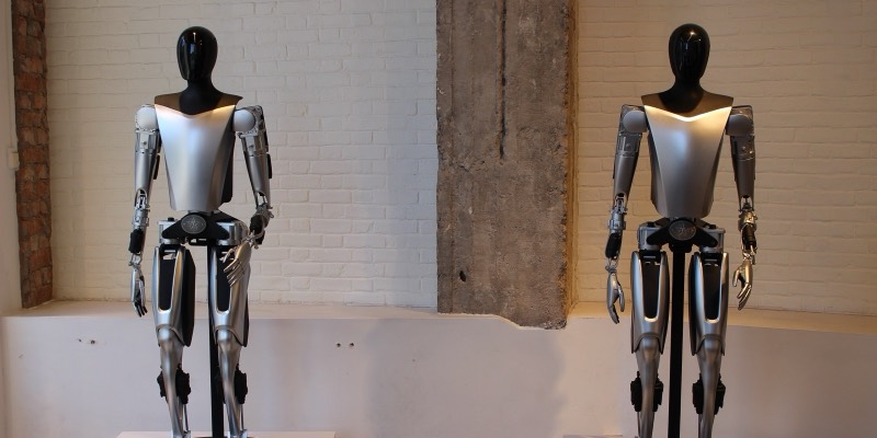 Two silver and black Tesla Optimus humanoid robots standing on display stands in front of a white brick wall