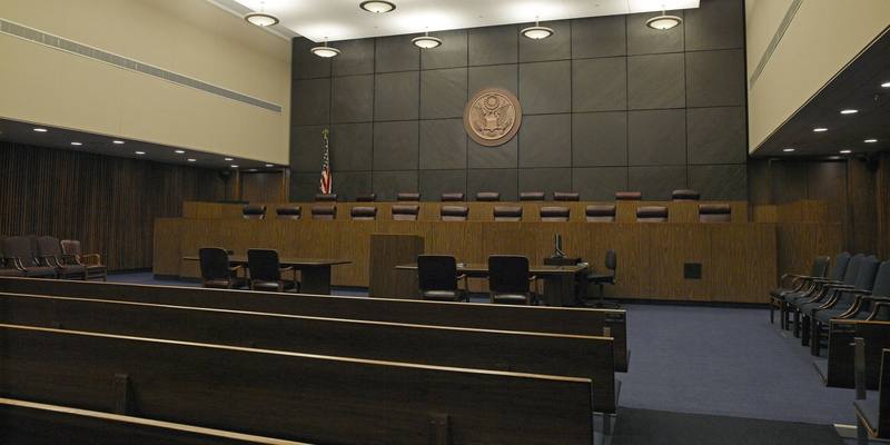 An empty wood-panelled courtroom with a long raised judges' bench, the US seal on a dark wall and rows of public benches in front