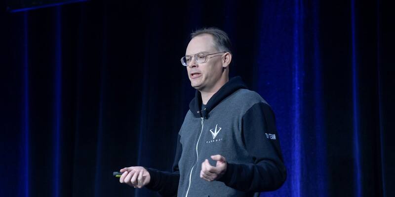 Tim Sweeney in glasses and a grey zip-up hoodie speaking on stage in front of a dark blue curtain