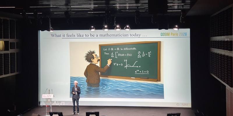 Mathematician Timothy Gowers speaks on a dark stage in front of a large screen showing a cartoon of a mathematician writing on a blackboard while standing waist-deep in rising water.