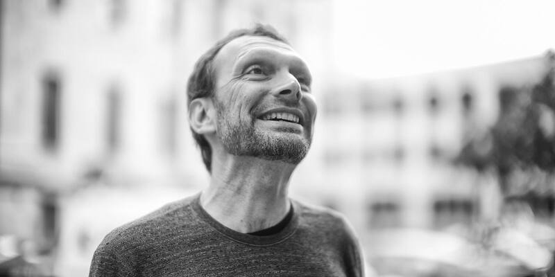Black and white photo of Toni Schneider, smiling and looking up, in a dark crew-neck jumper with blurred buildings behind him