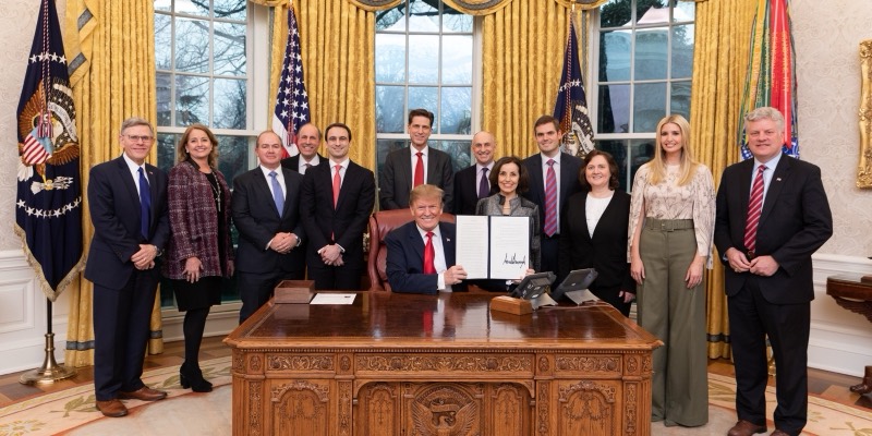 Donald Trump seated at the Resolute Desk holding up a signed executive order, surrounded by officials in the Oval Office
