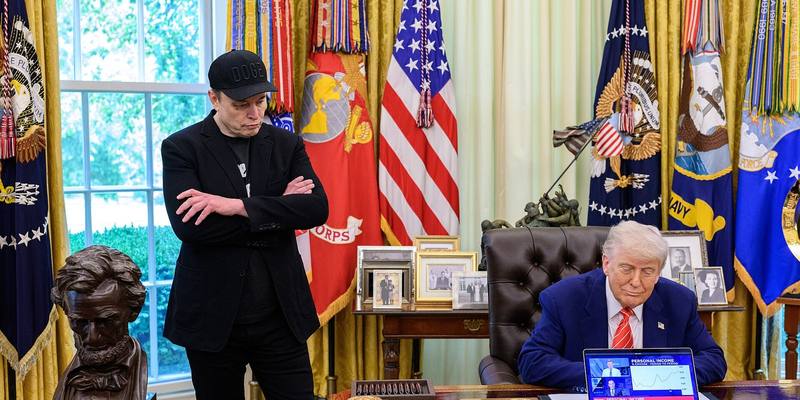 Elon Musk, arms folded and wearing a black cap, stands in the Oval Office as Donald Trump sits at the Resolute Desk in front of a laptop