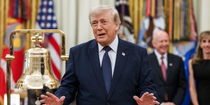 Donald Trump in a navy suit gesturing beside a brass New York Stock Exchange bell in the Oval Office