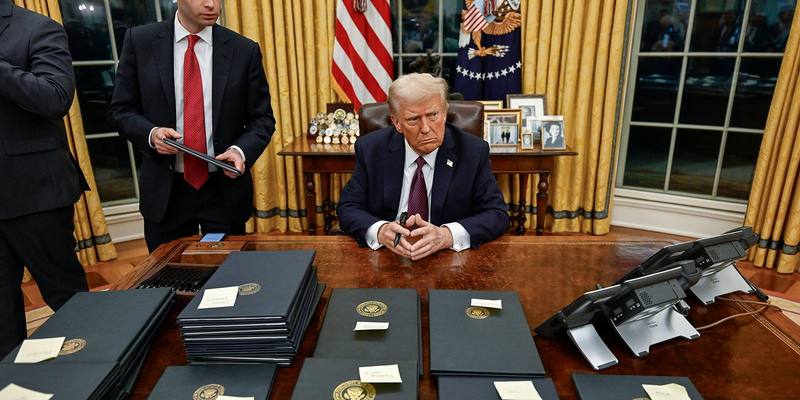 Donald Trump seated at the Resolute Desk behind rows of black executive order folders, with an aide holding a folder beside him and US flags behind
