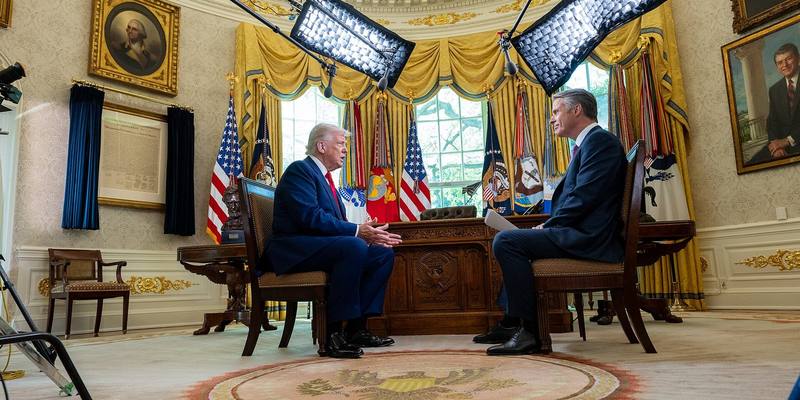Donald Trump seated in the Oval Office facing an interviewer, with flags, studio lights and the Resolute Desk behind them