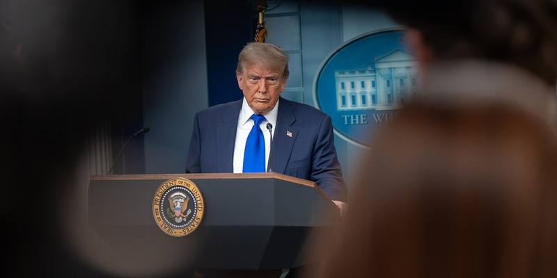 Donald Trump in a dark suit and blue tie standing at the presidential lectern in the White House press briefing room, framed between blurred reporters in the foreground