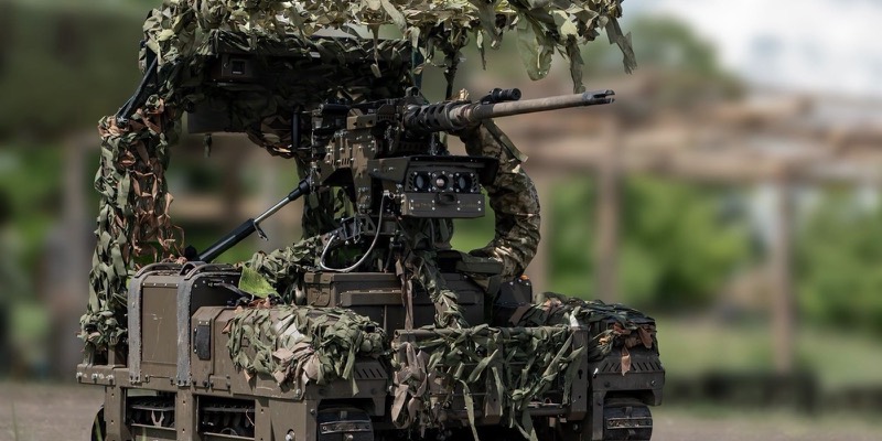 A tracked ground robot draped in camouflage netting, with a mounted heavy machine gun and a sensor camera, on a dirt training ground