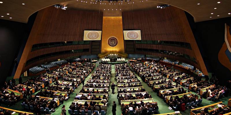 The UN General Assembly Hall seen from the back, with rows of delegates at green desks facing the gold wall and UN emblem at the front