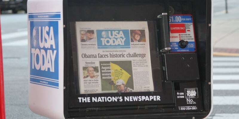 A white and black USA Today street vending box with a front page displayed behind its glass door and the words The Nation’s Newspaper beneath