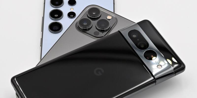 The camera bumps of three smartphones stacked on a white table: a pale blue Samsung Galaxy, a graphite iPhone Pro and a black Google Pixel on top