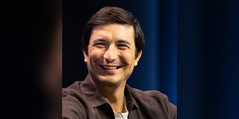 Robinhood CEO Vlad Tenev, with short dark hair and a brown shirt, smiling broadly against a dark blue background.
