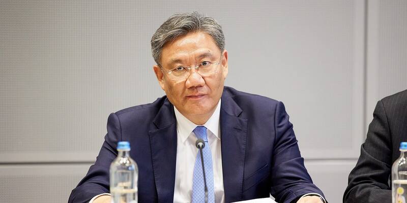 Wang Wentao in glasses, a navy suit and a light blue tie, seated at a conference table behind a microphone and a water bottle