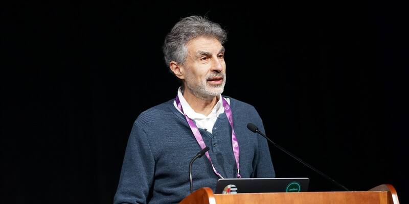 Yoshua Bengio, grey-haired with a beard, in a blue jumper and lanyard, speaking at a wooden podium against a black background