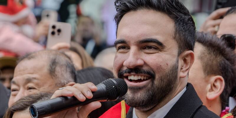 Zohran Mamdani smiling and speaking into a microphone in a crowd at a street celebration